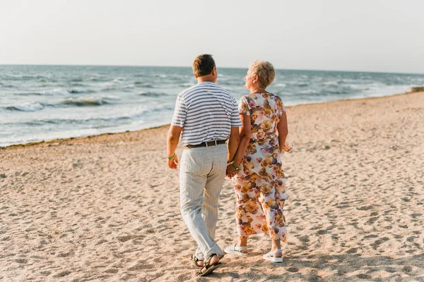 Sahilde birlikte duran romantik yetişkin çift. Olgun çiftler tatilde deniz kenarında dinlenirler. Mutlu aile yürüyüşleri ve deniz kenarında el ele tutuşmalar. Karı koca kucaklaşması. Okyanusa yakın bir aşk hikayesi.