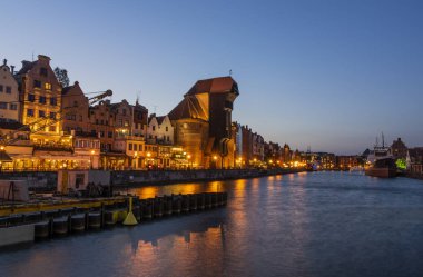 Gün batımında Motlawa Nehri kıyısında Gdansk Eski şehir. Binalar arasında ünlü ortaçağ vinç. Ön planda bazı tekneler ve inşaat ekipmanları