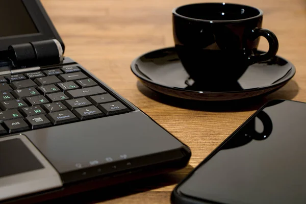 Remote workplace. Remote work environment. View of a natural wood table with several items, a black laptop, a cup of coffee and a mobile phone.