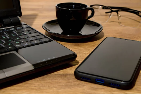 Remote workplace. Remote work environment. View of a natural wood table with several items, a black laptop, glasses, a cup of coffee and a mobile phone.