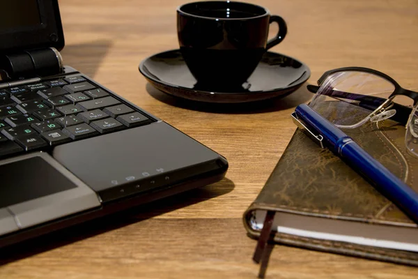 Remote workplace. Remote work environment. View of a natural wood table with several items, a black laptop, glasses, a cup of coffee, a diary and a pen.