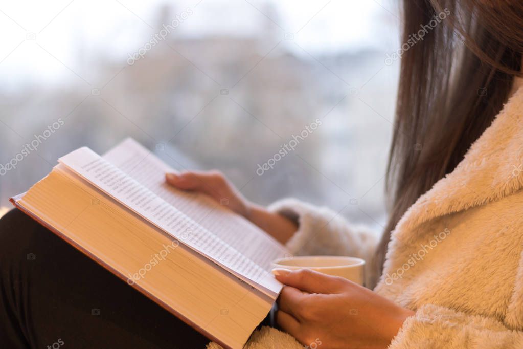 cropped shot of woman reading book with cup of hot beverage while relaxing at home in front of window