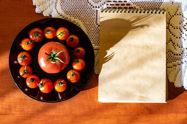厨房桌子上的黑盘子里的红色西红柿 旁边是一个记事本 — 图库照片