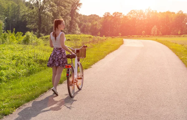 Girl with bike at the summer sunset on the road in the city park. Cycle closeup wheel on blurred summer background. Cycling down the street to work at summer sunset. Bicycle and ecology lifestyle concept.