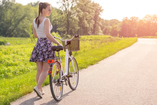 Girl with bike at the summer sunset on the road in the city park. Cycle closeup wheel on blurred summer background. Cycling down the street to work at summer sunset. Bicycle and ecology lifestyle concept.