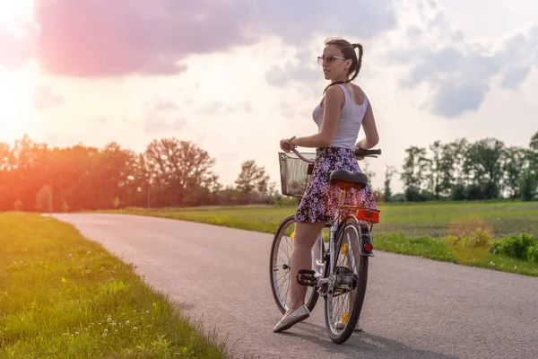 Girl with bike at the summer sunset on the road in the city park. Cycle closeup wheel on blurred summer background. Cycling down the street to work at summer sunset. Bicycle and ecology lifestyle concept.