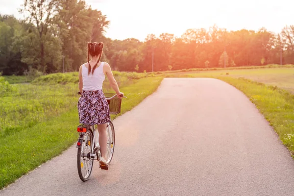 Girl close up with bike at the summer sunset on the road in the city park. Cycling down the street to work at summer sunset. Bicycle and ecology lifestyle concept.