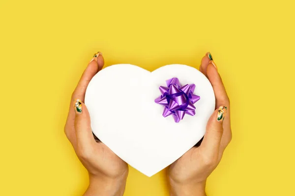 Heart shaped Valentines Day gift box in the hands of the girl on yellow paper background.