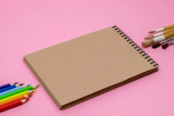 色鉛筆、水彩画ブラシ、モックアップ用のピンクの背景に隔離されたノートブック。フラットレイ、トップビュー。学校の概念に戻る. — ストック写真
