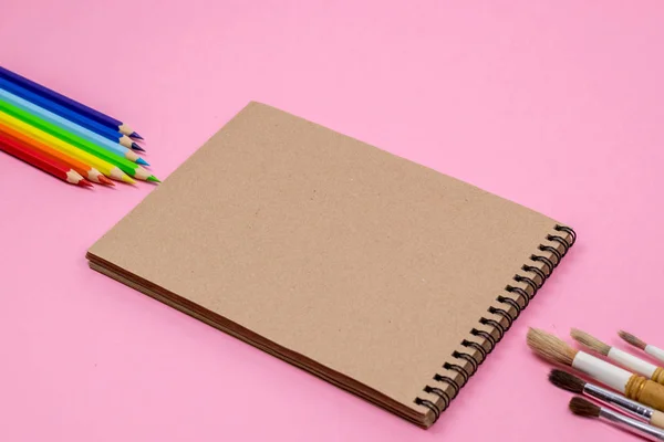 色鉛筆、水彩画ブラシ、モックアップ用のピンクの背景に隔離されたノートブック。フラットレイ、トップビュー。学校の概念に戻る. — ストック写真