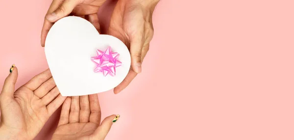 Widok z góry męskich i żeńskich rąk posiadających kształt serca pudełko z różowym kokardą na różowym tle płaski lay. Prezent na urodziny, Walentynki, Boże Narodzenie, nowy rok. — Zdjęcie stockowe
