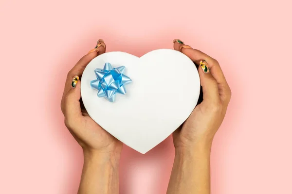 Kobiece ręce trzymać kształt serca czerwony prezent z niebieskim łuku na różowym tle. Jest wolne miejsce na tekst. — Zdjęcie stockowe