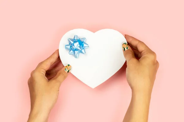 Las manos femeninas sostienen un regalo en forma de corazón rojo con un lazo azul sobre un fondo rosa. Hay espacio libre para el texto . —  Fotos de Stock