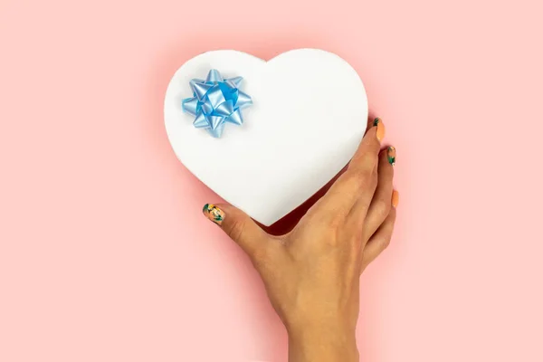 Las manos femeninas sostienen un regalo en forma de corazón rojo con un lazo azul sobre un fondo rosa. Hay espacio libre para el texto . — Foto de Stock