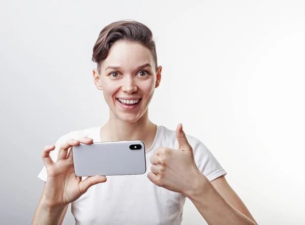 Menina adolescente lindo está tirando uma foto usando câmera frontal em seu smartphone e mostrando o polegar para cima. Isolado sobre fundo branco . — Fotografia de Stock