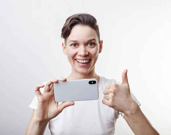 Menina adolescente lindo está tirando uma foto usando câmera frontal em seu smartphone e mostrando o polegar para cima. Isolado sobre fundo branco . — Fotografia de Stock