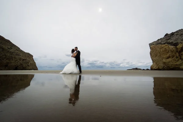 The bride and groom walk along the beach, reflecting in the water. Panoramic view of reflection. Walking along the beach against the rocks and ocean waves.