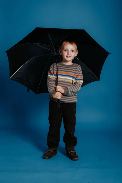 Un niño pequeño está de pie bajo un paraguas abierto y sonriendo . — Foto de Stock