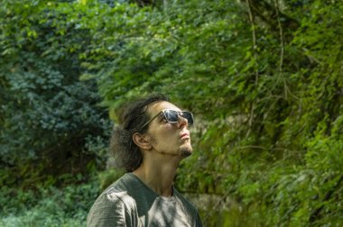 Dışarı çık. Güneş gözlüklü genç bir adam çevresindeki doğaya bakar. Gün ışığı yüzüne vurur.