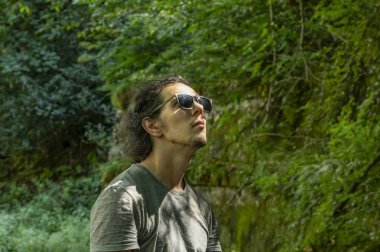 Dışarı çık. Güneş gözlüklü genç bir adam çevresindeki doğaya bakar. Gün ışığı yüzüne vurur.