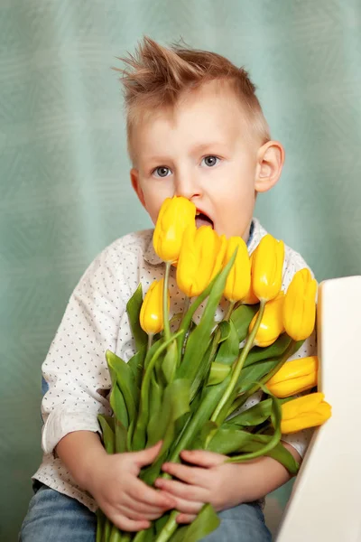 Adorable smiling child with spring flower bouquet looking at camera isolated on blue. Little toddler boy holding yellow tulips as gift for mom