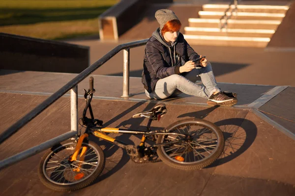 女の子は夕日のヘルメットのアスファルトの上に自転車スタンドに座っています — ストック写真