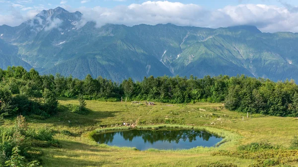 View of a mountain glade with a transparent mirror lake, and tourists and high mountains with snow-capped peaks in the distance. Caucasus, Russia