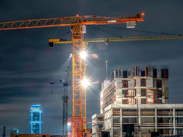 Tower cranes at a construction site in the night city. Electric light with rays.