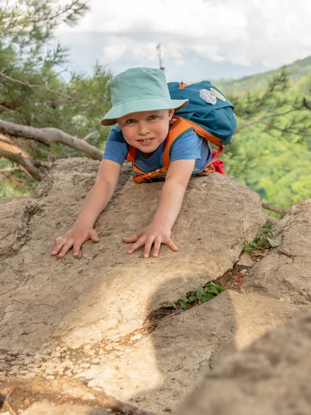 A boy traveler with a backpack climbed to the top of a cliff surrounded by a green forest.