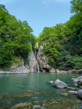 Yüksek bir şelale uçurumdan açık bir göle düşüyor. Şelale yeşil ormanıyla çevrilidir