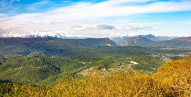 Ufukta evler ve yüksek karlı dağlar ile yeşil bir tepelik vadide gün batımında görünümü.