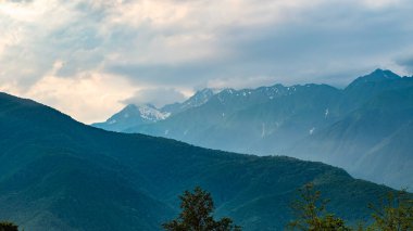 Yeşil ve kayalık yamaçları ile dağ sıraları. Bulutların içinde karlı zirveleri olan dağlar.