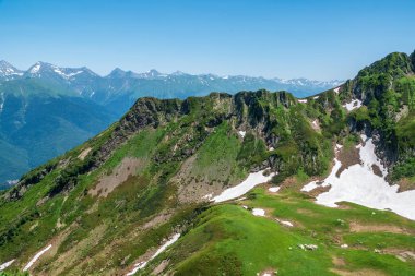 Açık mavi gökyüzüne karşı yamaçlarda kayalar ve kar ile dağ sırası.