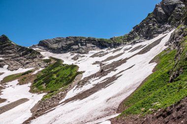 Açık mavi gökyüzüne karşı yamaçlarda kayalar ve kar ile dağ sırası.
