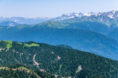 Yüksek dağlarla çevrili yeşil dağ vadisine yükseklikten görünümü. Ufukta karla kaplı dağ zirveleri. Krasnaya Polyana, Soçi, Kafkasya, Rusya Federasyonu.