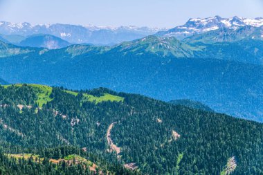 Yüksek dağlarla çevrili yeşil dağ vadisine yükseklikten görünümü. Ufukta karla kaplı dağ zirveleri. Vadide bir tepede Yalnız ev.