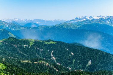 Yüksek dağlarla çevrili yeşil dağ vadisine yükseklikten görünümü. Ufukta karla kaplı dağ zirveleri. Krasnaya Polyana, Soçi, Kafkasya, Rusya Federasyonu.