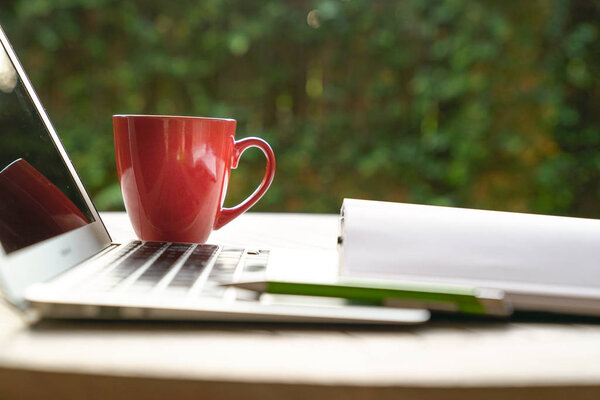 Work from home concept: laptop, red mug and note pad on a table with focus on a green pen with garden background