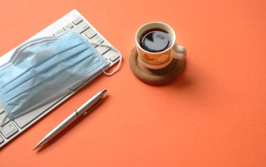Klavyeli parlak turuncu masa, tıbbi yüz maskesi, kalem ve kahve..