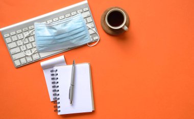 Klavyeli parlak turuncu masa, tıbbi maske, kahve ve not defteri. Üst görünüm.