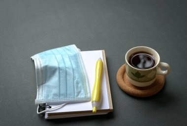 Tıbbi yüz maskeli kahve fincanı, not defteri ve kırsal siyah masa üstünde kalem. Boşluğu kopyala.