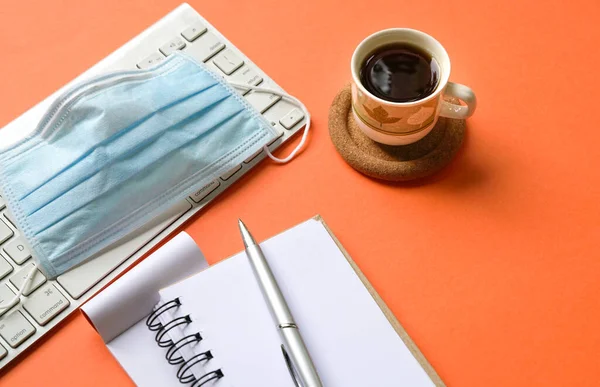 Klavyeli parlak turuncu masa, tıbbi maske, kahve ve not defteri. Üst görünüm.