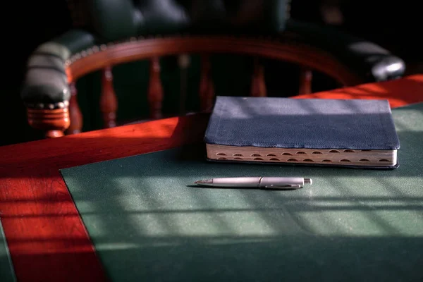 Closed bible on the edge of table with chesterfield chair at the background. Light play forming lines and patterns on surface.