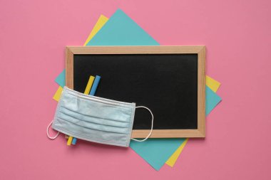 Siyah tahtanın üstünde yüz maskesi, renkli kalem ve kağıtlar. Pembe arkaplan, üst görünüm.