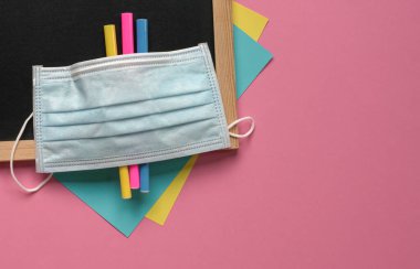 Siyah tahtanın üstünde yüz maskesi, boyama kalemleri ve kağıtlar. Pembe arka plan, üst manzara. Boşluğu kopyala.