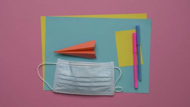 Kalemlerin üstüne yüz maskesi ve kağıt uçakla kağıtlar. Pembe arkaplan.