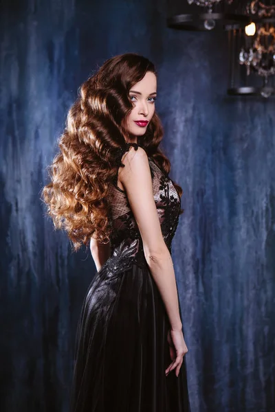 Young woman with long curly hair and makeup in evening long luxury dress, posing in a dark interior room. fashion beauty portrait