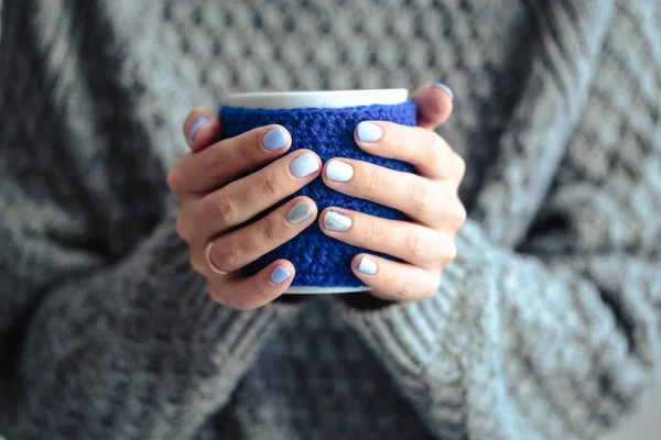 Gorgeous manicure, pastel tender color nail polish, closeup photo. Female hands hold a cup in a knit blue cover