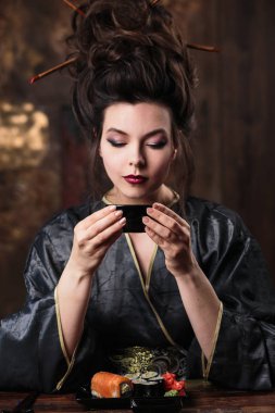 Bir geyşa Asya kostüm moda makyaj ve saç stili ile şehvetli genç kadın çay içiyor ve suşi, moda güzellik portre yiyor