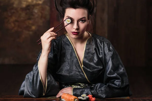 Sensual young woman in a geisha asian costume with fashion makeup and hair style eats sushi, fashion beauty portrait